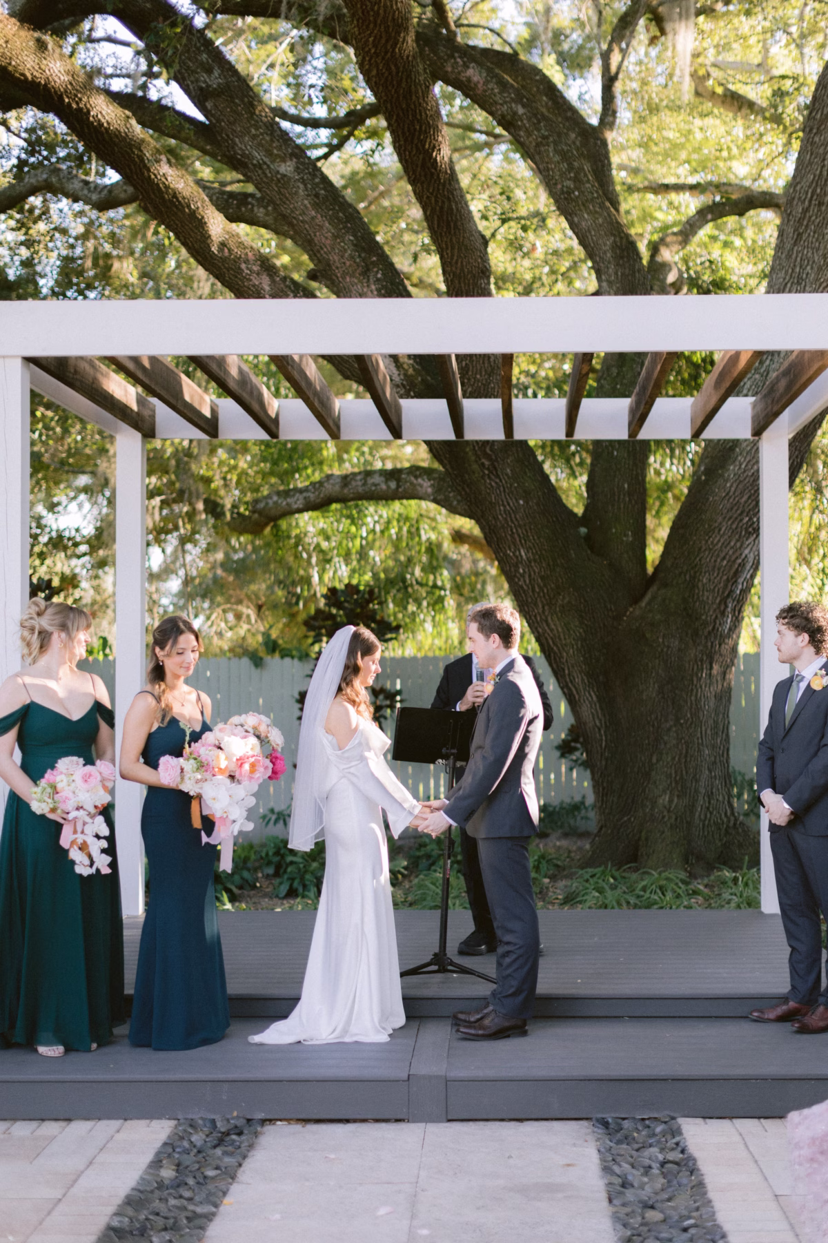 Outdoor wedding ceremony at Soire Estate in Tampa featuring bridesmaids in emerald green dresses holding bouquets by Golden Wild Florals under a modern pergola