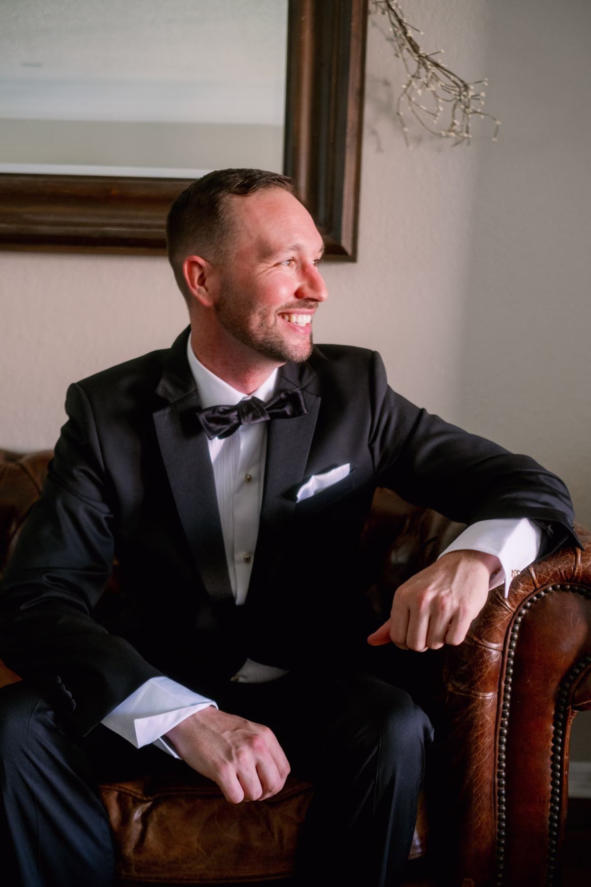 Elegant groom in black tuxedo from Nordstrom sitting in leather chair during formal wedding portrait session at The Vinoy St. Petersburg