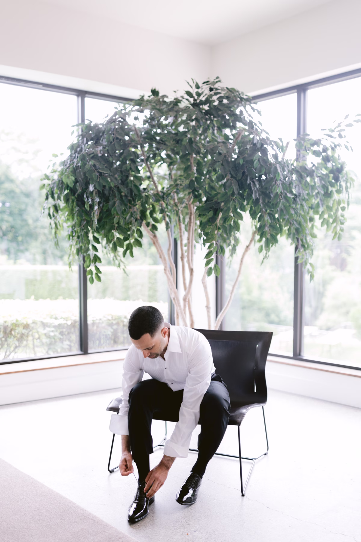 Groom putting on dress shoes in modern corner room with large windows and greenery at The Roundhouse Beacon Hudson Valley wedding venue