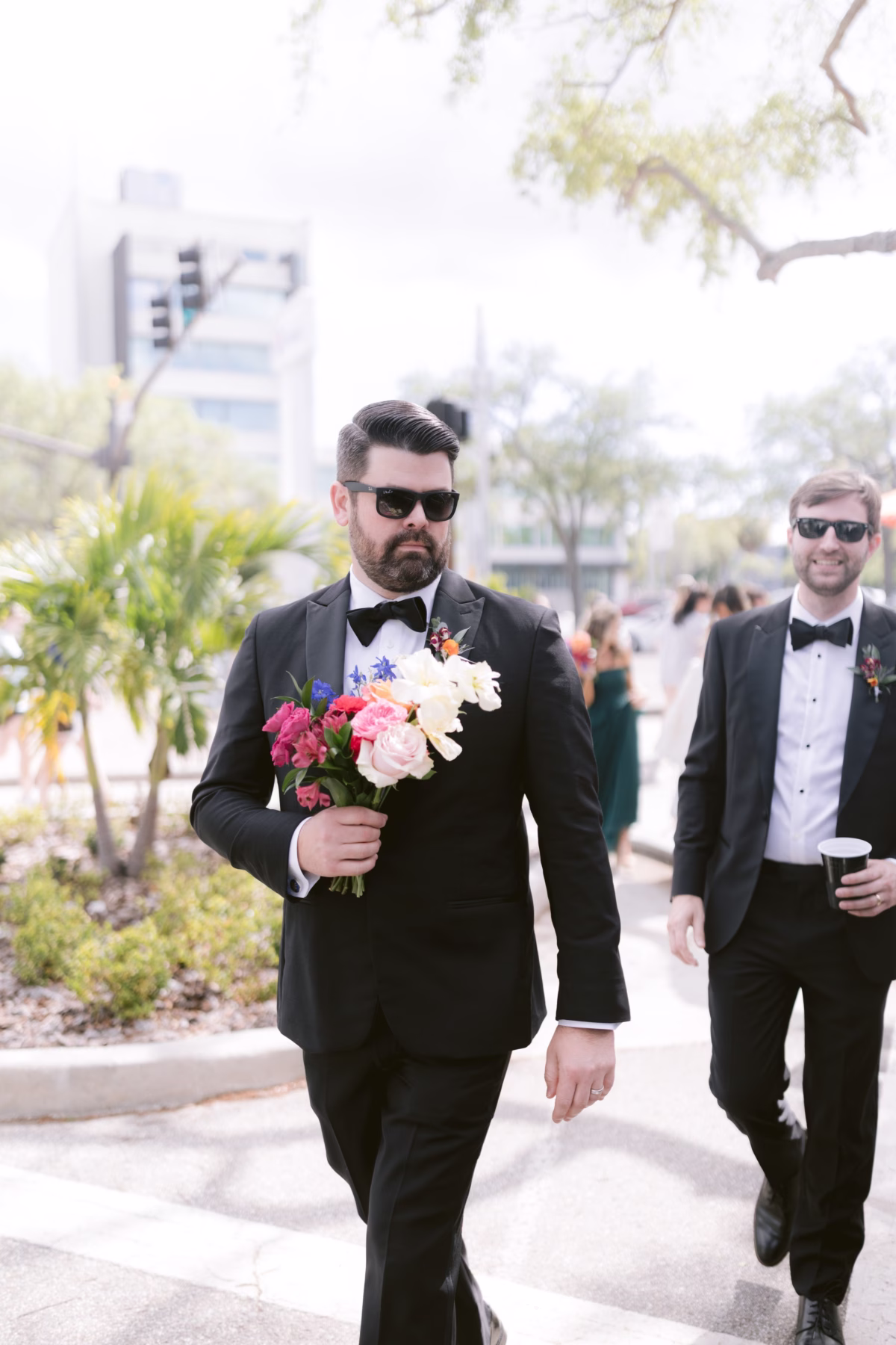 Groom in black tuxedo carrying colorful bouquet walks with groomsman at St Pete Shuffleboard Club wedding with florals by Petite Fleur