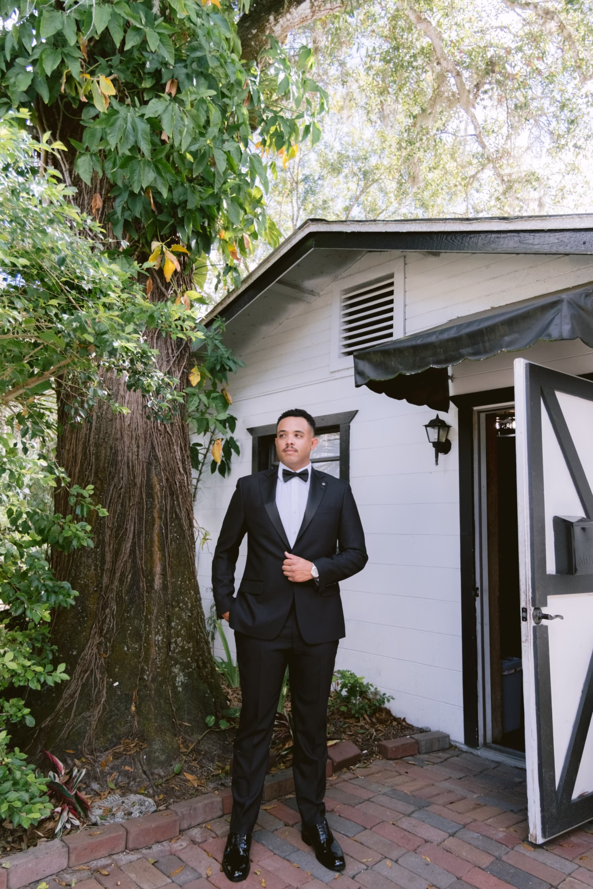 Elegant groom in black tuxedo posing outside charming cottage at Soire Estate Tampa wedding planned by Daniela Alvarez Event Planning