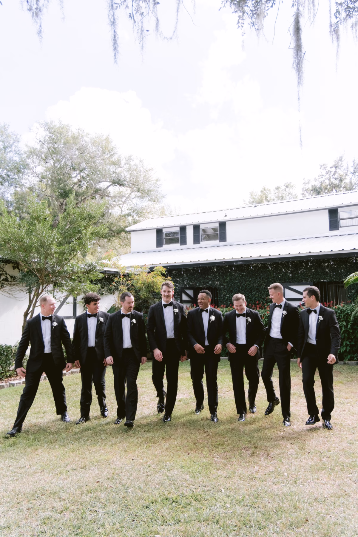 Eight groomsmen in classic black tuxedos with bow ties pose together on the lawn at Soire Estate Tampa wedding photographed by Ashley Izquierdo Photography