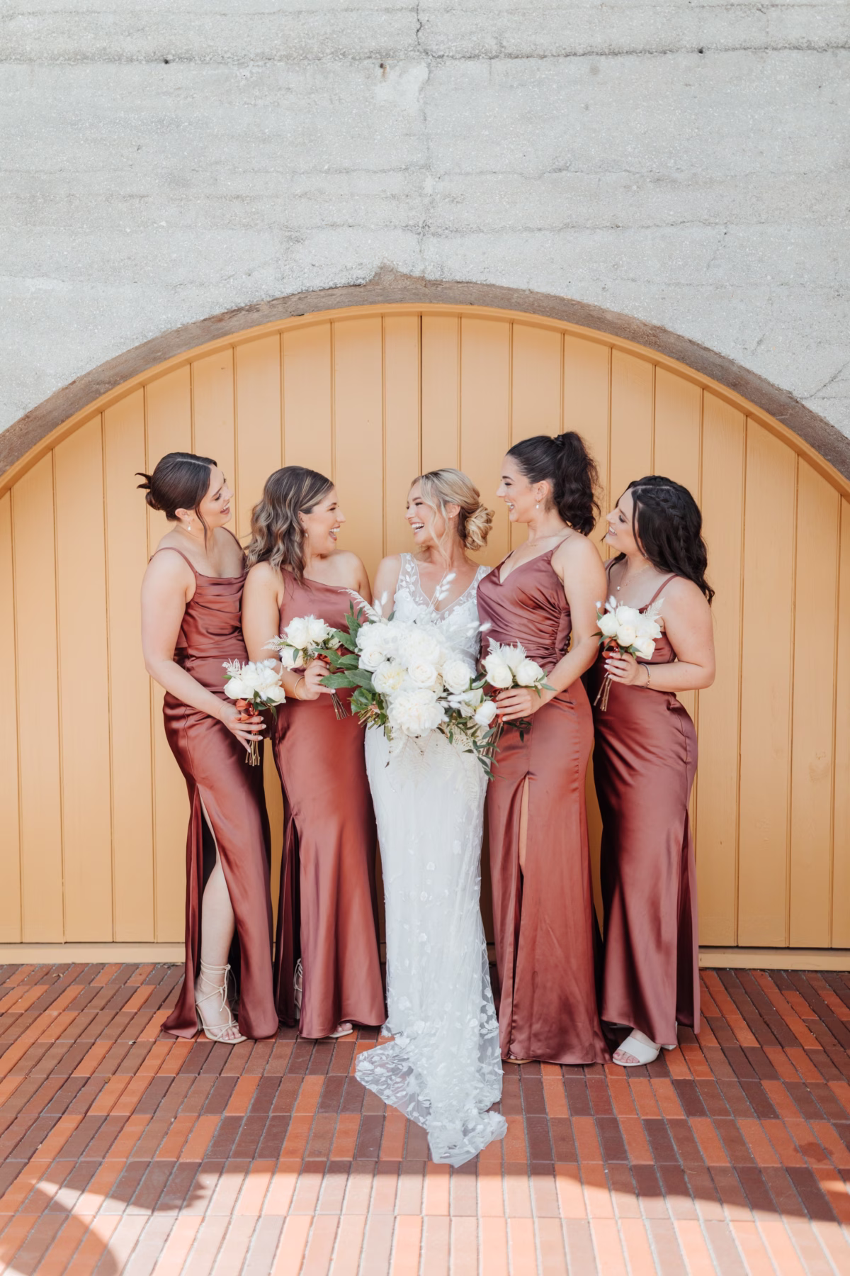 Bride in Alexandra Grecco gown with bridesmaids in rust dresses holding white bouquets by Aime Peterson Flowers at The Lightner Museum historic wedding venue