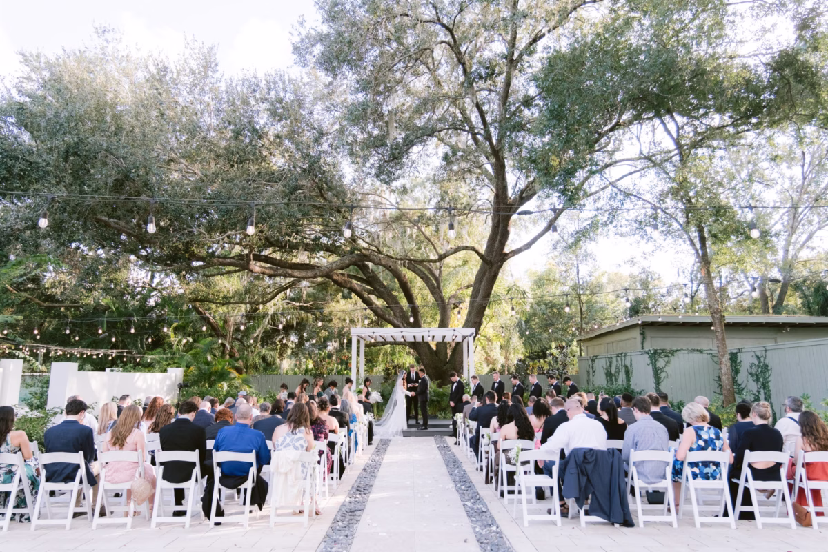 Outdoor wedding ceremony at Soire Estate in Tampa with white chairs lined along aisle under large oak tree with string lights, planned by Daniela Alvarez Event Planning