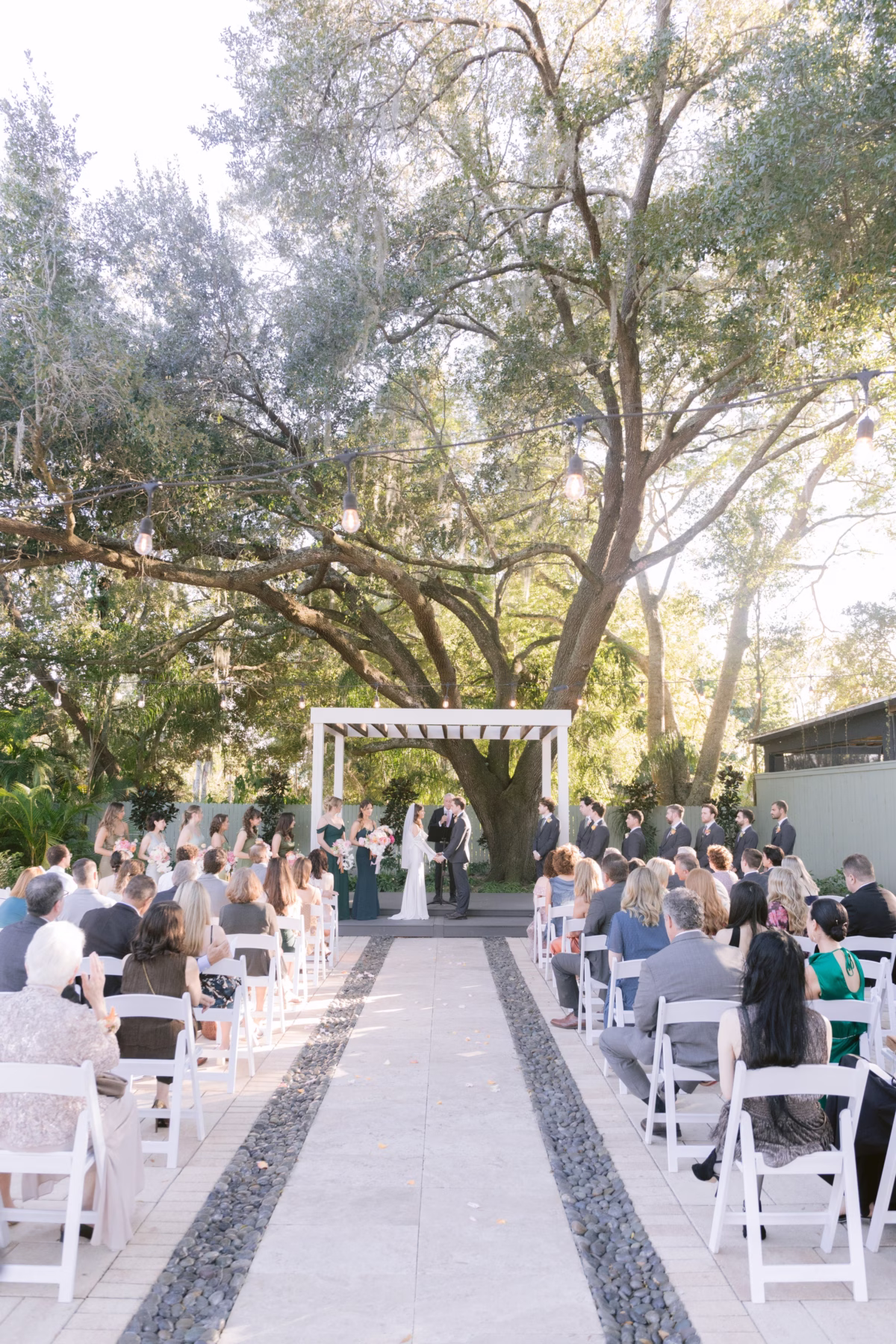 Outdoor wedding ceremony at Soire Estate in Tampa with guests seated in white chairs watching bride and groom exchange vows under white pergola surrounded by lush trees