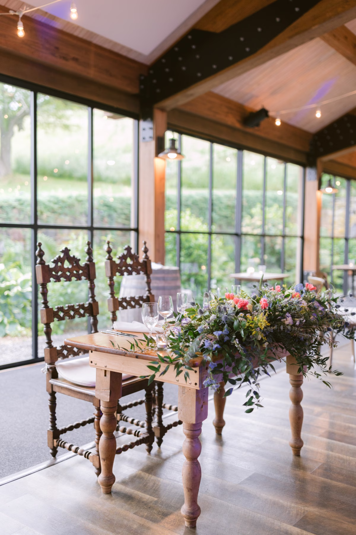 Elegant wedding reception tablescape with wildflower centerpiece by Cherry Brandy Designs at Red Maple Vineyard featuring vintage wooden chairs and floor-to-ceiling windows