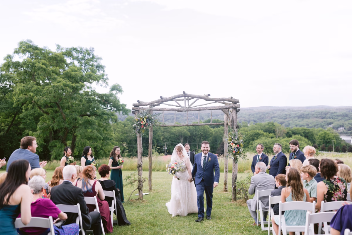 Newlyweds walk down the aisle after their outdoor wedding ceremony under a rustic wooden arbor with wildflower arrangements by Cherry Brandy Designs at Red Maple Vineyard