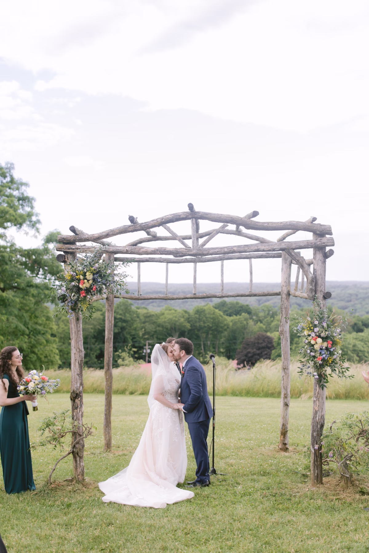 Bride and groom kiss under rustic wooden arbor with wildflower arrangements by Cherry Brandy Designs during outdoor wedding ceremony at Red Maple Vineyard