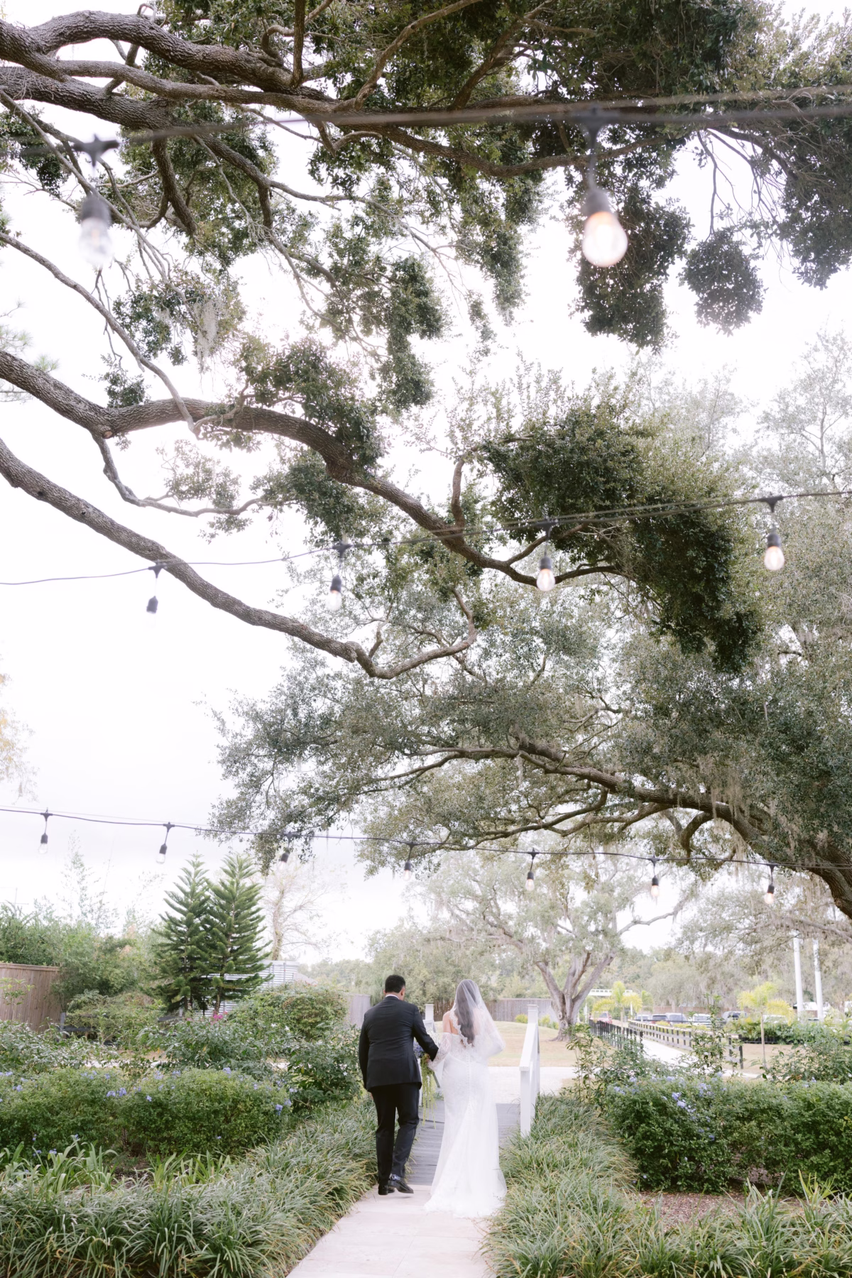 Bride and groom walking through garden pathway under string lights at Soire Estate Tampa wedding venue