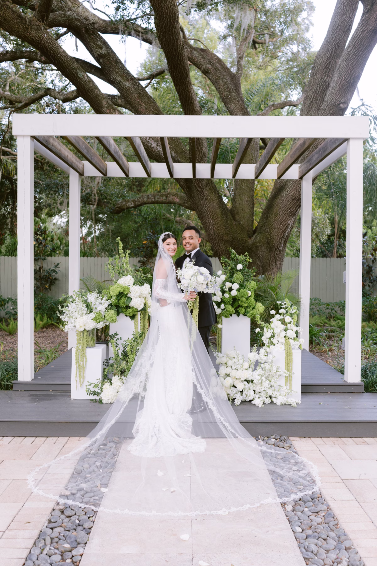 Bride and groom pose under white pergola with lush white and green floral arrangements by Ava Elise Decor at Soire Estate Tampa wedding