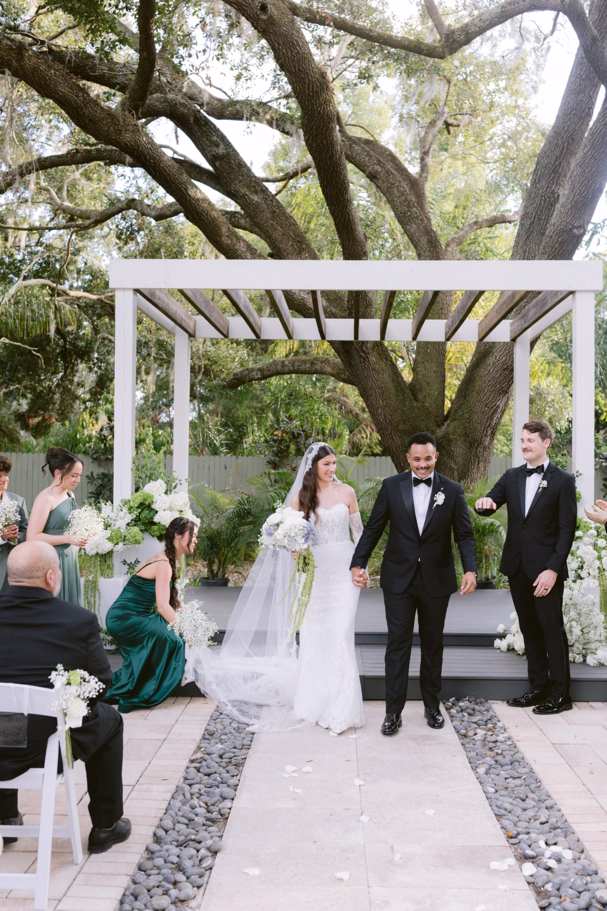 Bride and groom walking down aisle after wedding ceremony at Soire Estate in Tampa with white floral arrangements by Ava Elise Decor under large oak tree