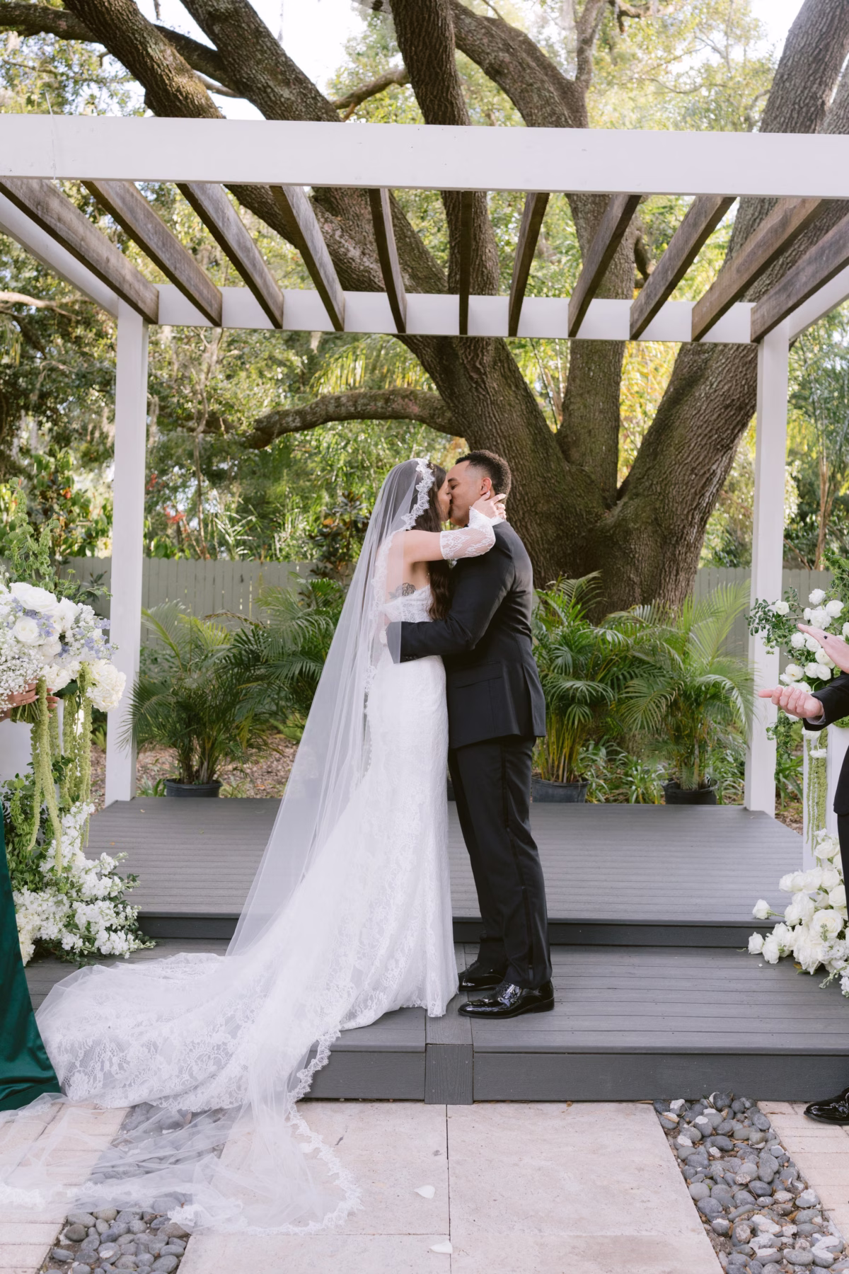 Bride and groom share first kiss under white pergola with white floral arrangements by Ava Elise Decor at Soire Estate Tampa wedding ceremony