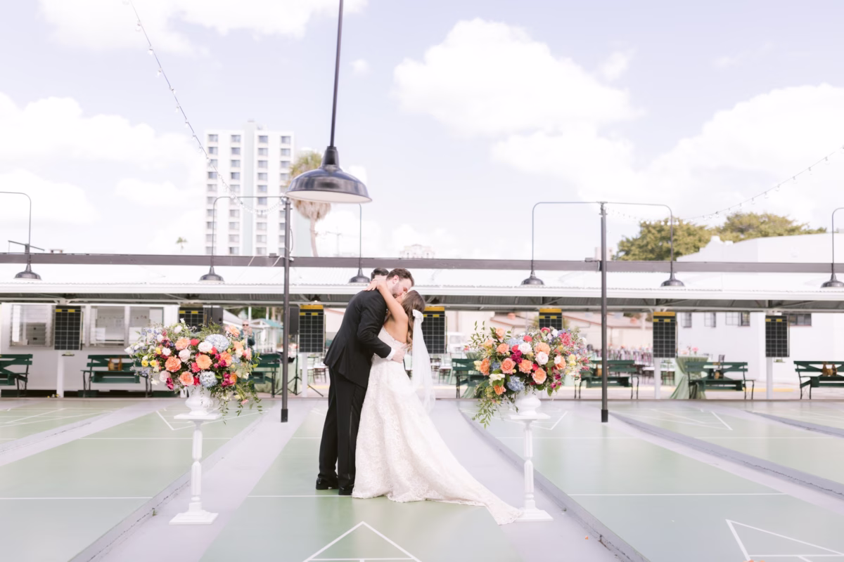 Bride and groom share their first kiss during wedding ceremony at St. Pete Shuffleboard Club with colorful floral arrangements by Petite Fleur