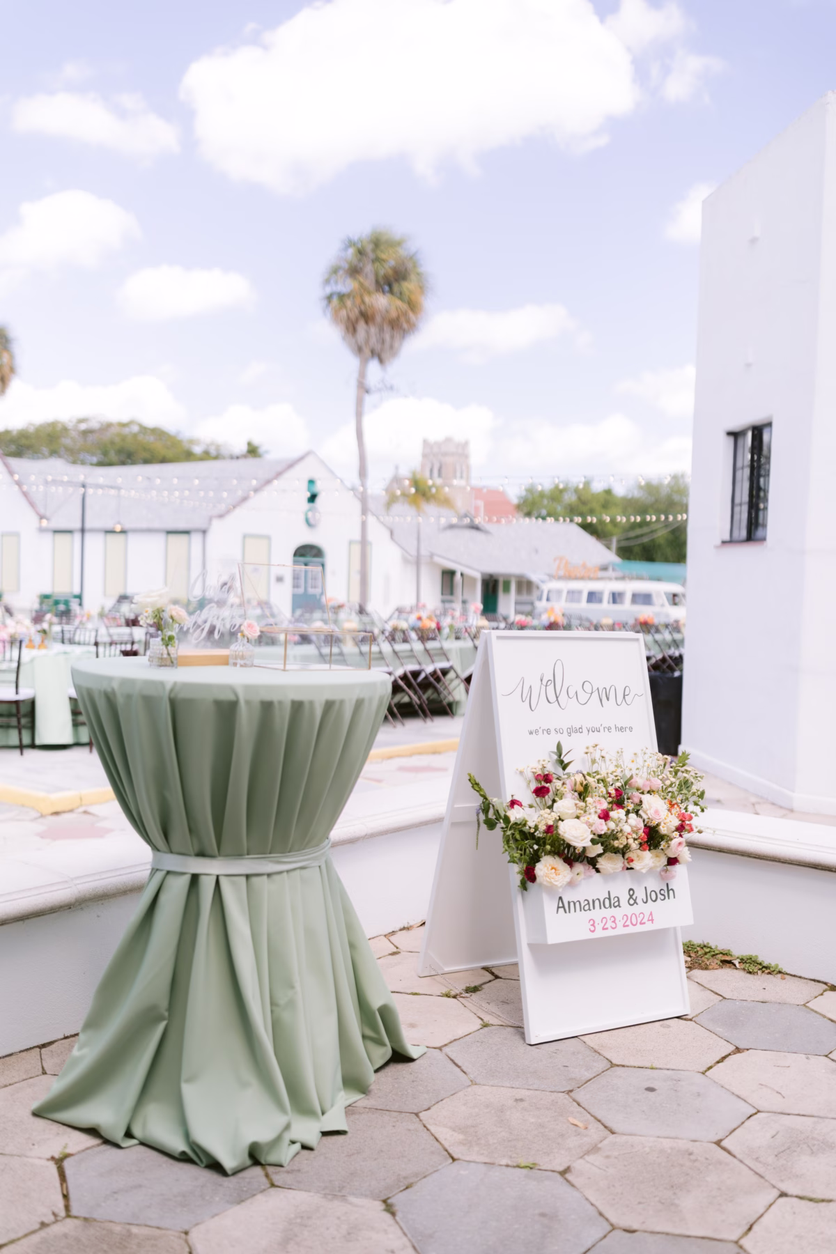 Sage green cocktail table and white welcome sign with floral arrangement by Petite Fleur at St Pete Shuffleboard Club wedding planned by Day of Details