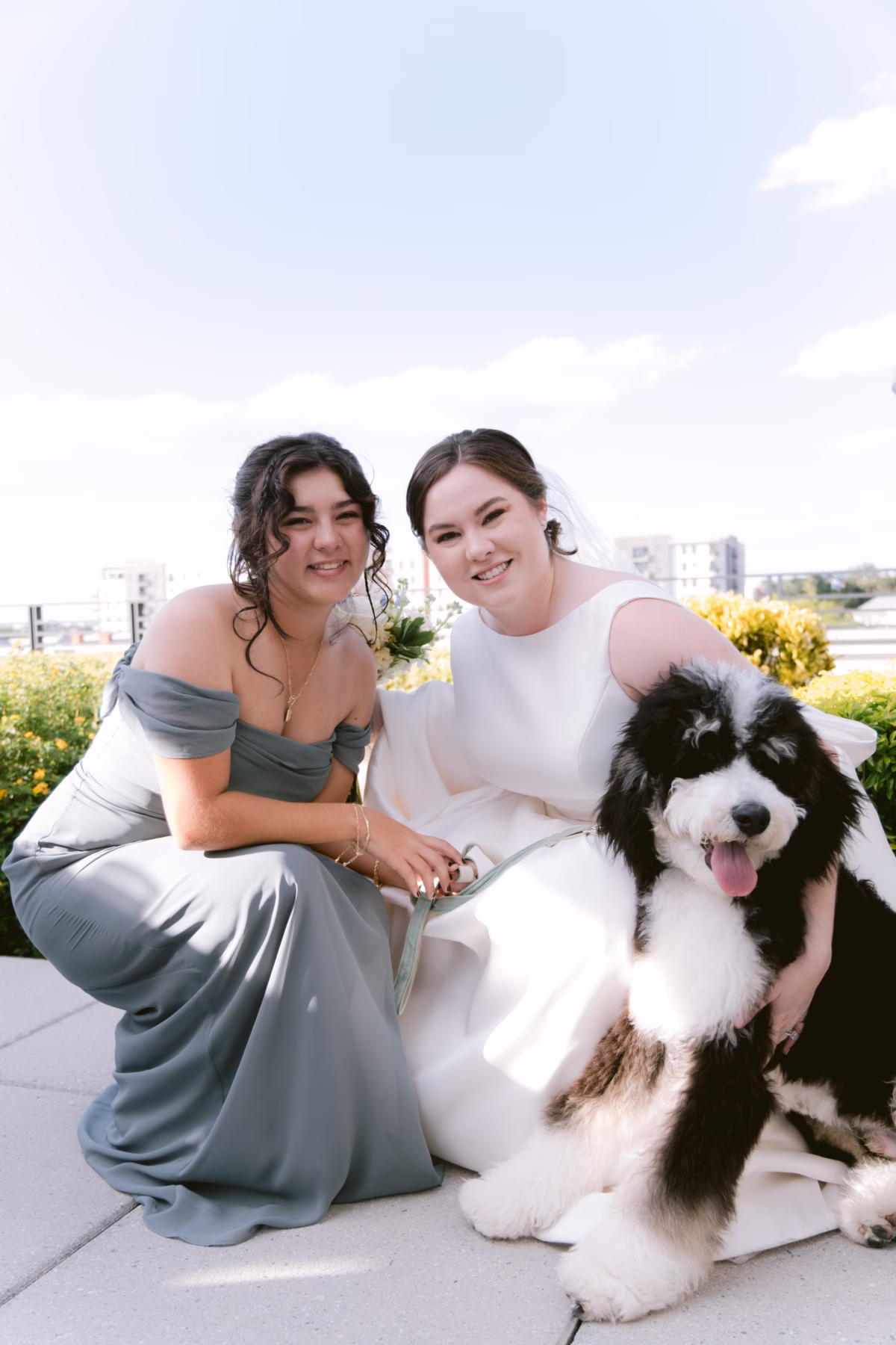 Bride in white dress and bridesmaid in sage green gown pose with fluffy black and white dog at Armature Works Tampa wedding planned by Logan Paradiso