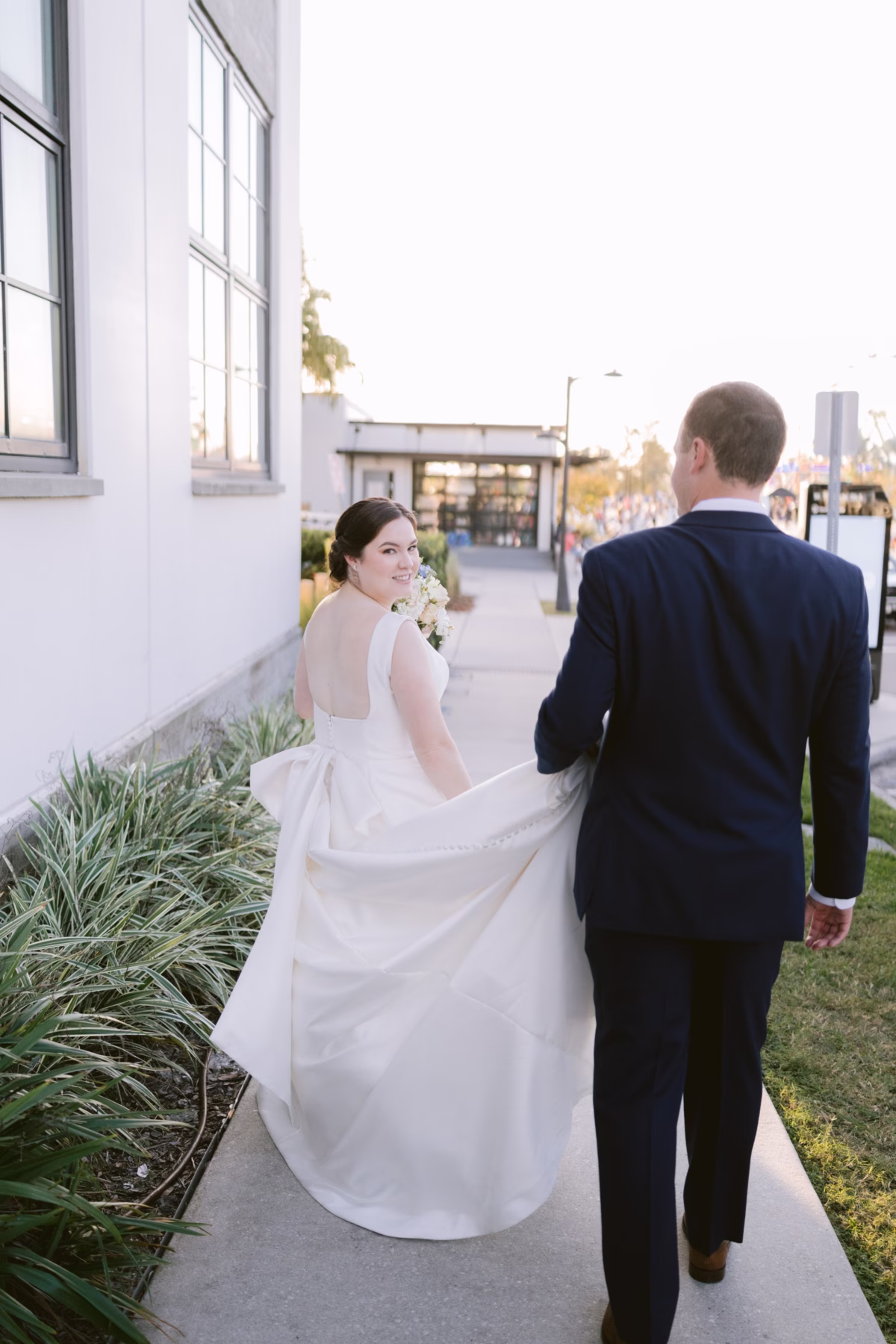Bride and groom walking together at Armature Works Tampa wedding planned by Logan Paradiso with bride wearing The White Closet Bridal gown and carrying I Dew Events white bouquet