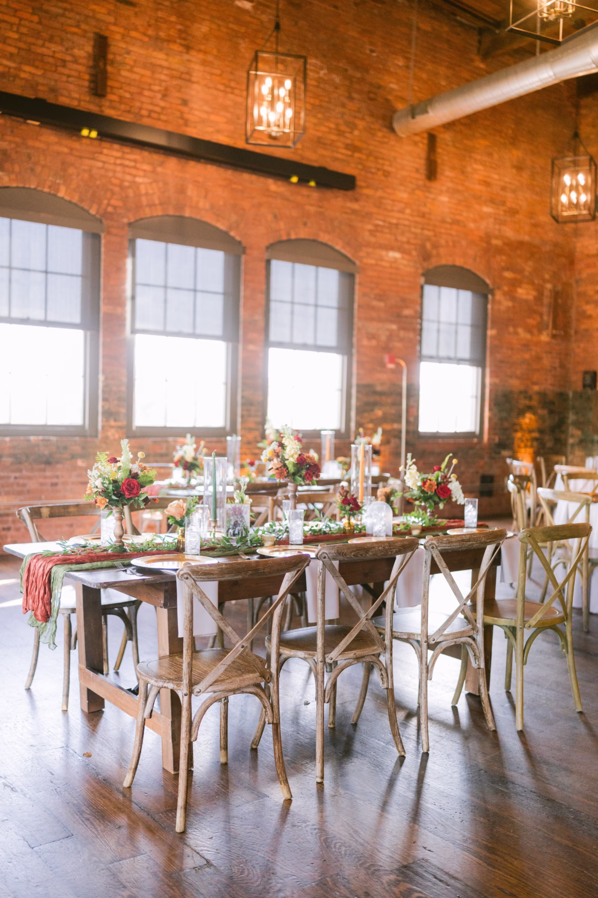 Rustic wooden wedding reception table with burgundy and blush florals by Events in Bloom at Armature Works Tampa featuring exposed brick walls and cross-back chairs