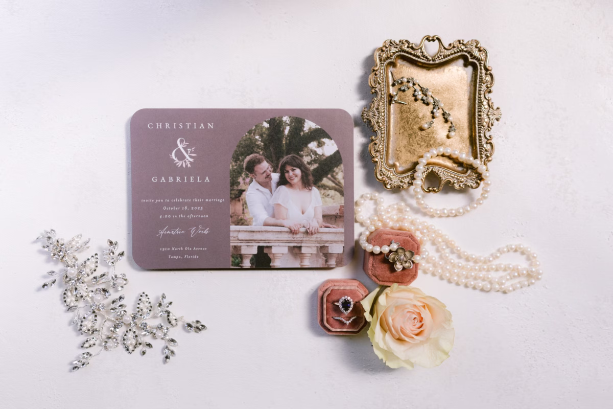 Wedding invitation flatlay featuring custom stationery by Seashine Weddings with engagement photo, vintage frame, pearl necklace, engagement ring, and blush rose for Armature Works Tampa wedding