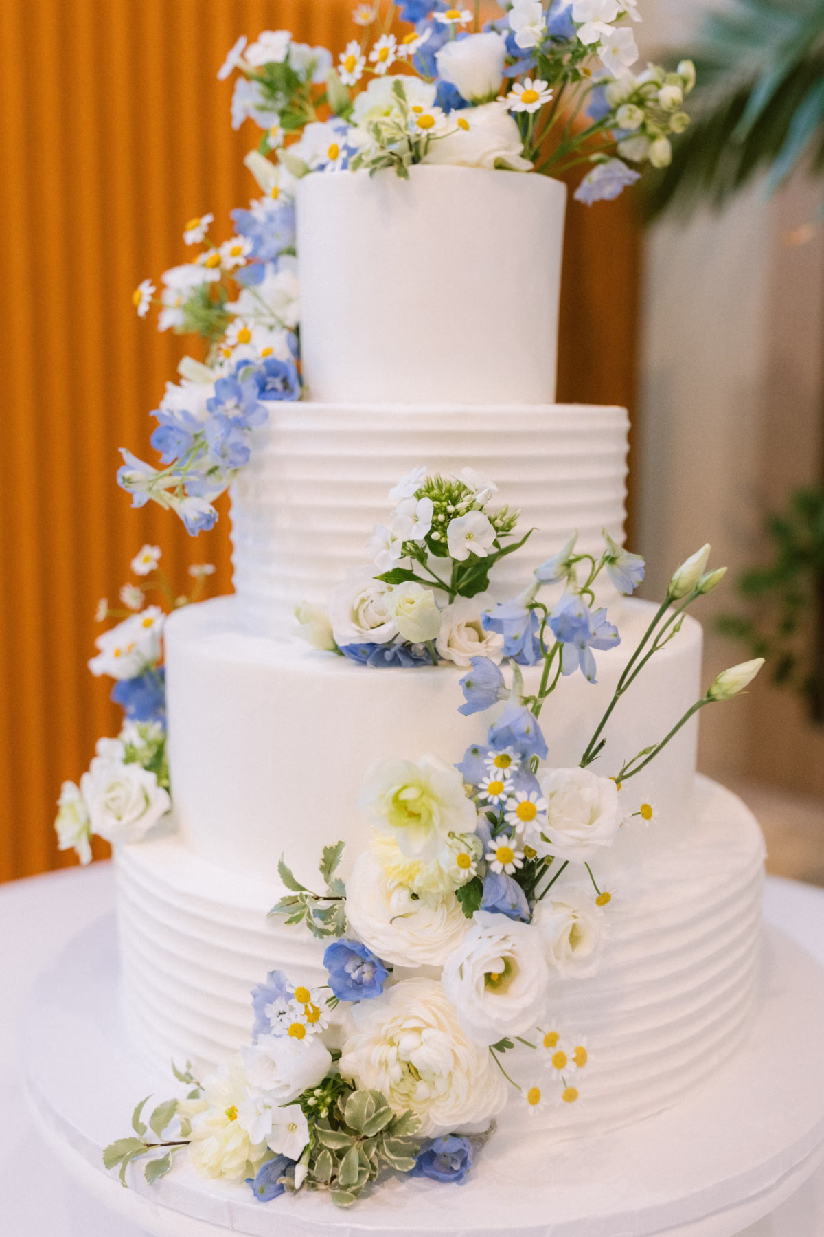 Elegant white four-tier wedding cake decorated with blue and white fresh flowers by Sweet Diva's Cakery and Wonderland Floral Art at St. Petersburg art museum wedding