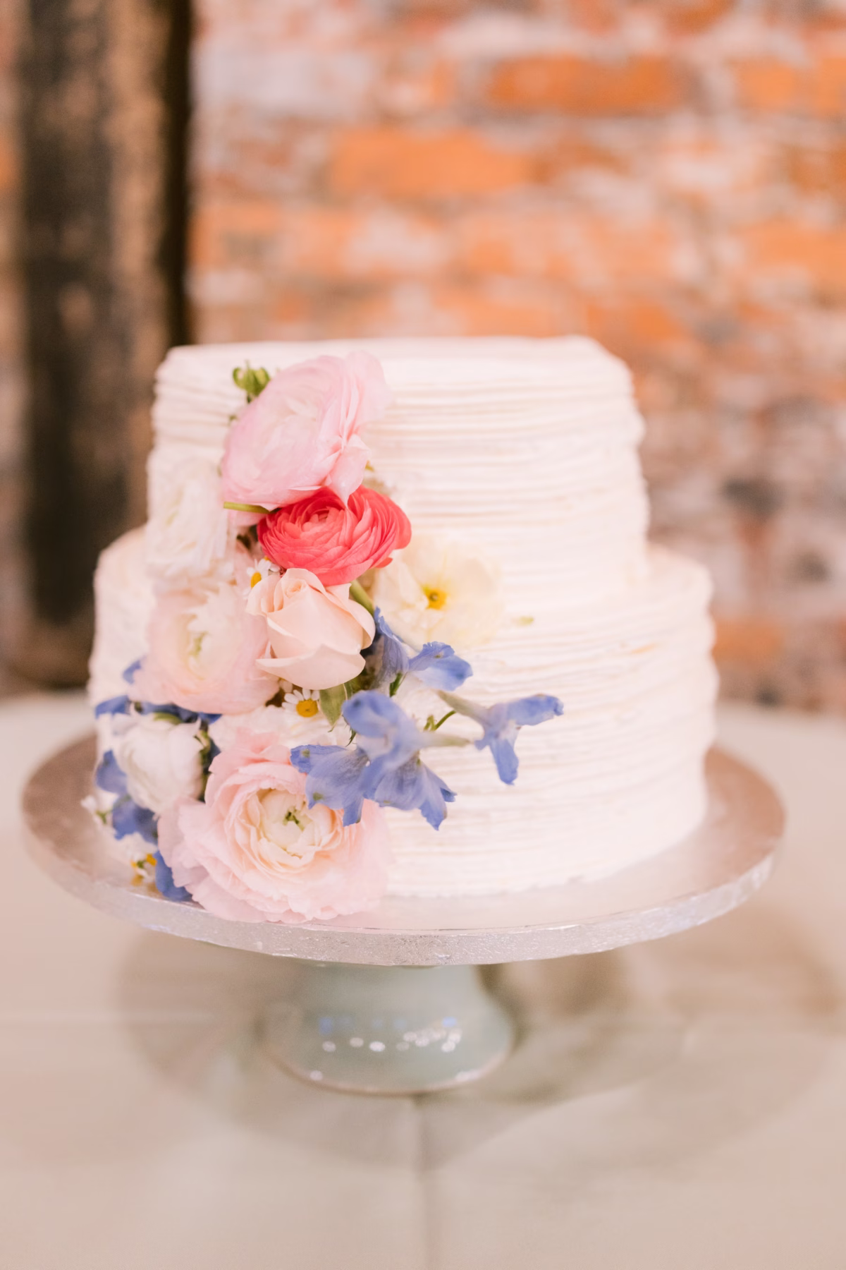 Two-tier white wedding cake decorated with pink ranunculus and blue flowers by I Dew Events at Armature Works Tampa wedding photographed by Ashley Izquierdo Photography